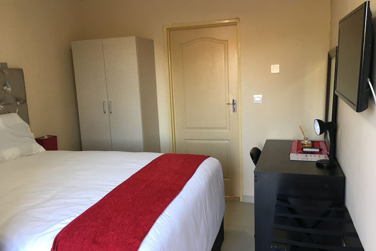 A hotel room with a bed, a wardrobe, a desk with a lamp, and a mirror.