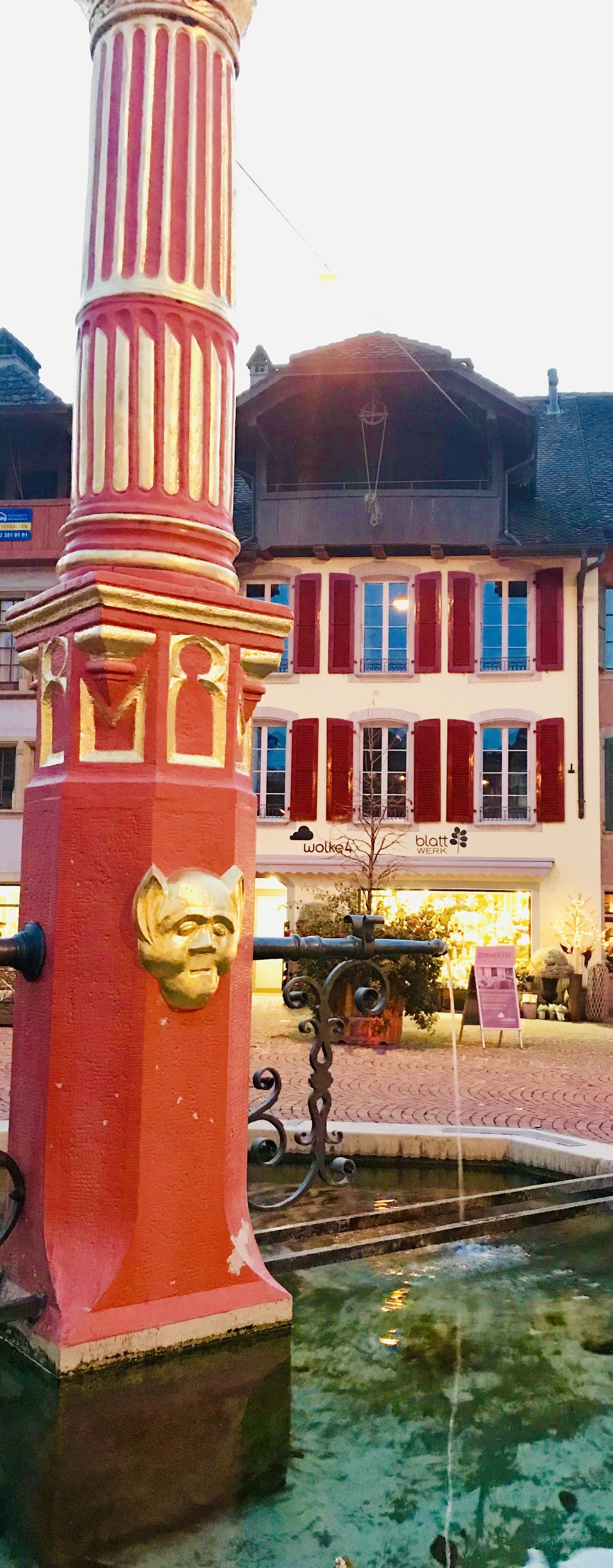 Une colonne rouge et blanche ornée d’un visage, une fontaine et un bâtiment aux volets rouges et à l’enseigne portant l’inscription « blatt WERK ».