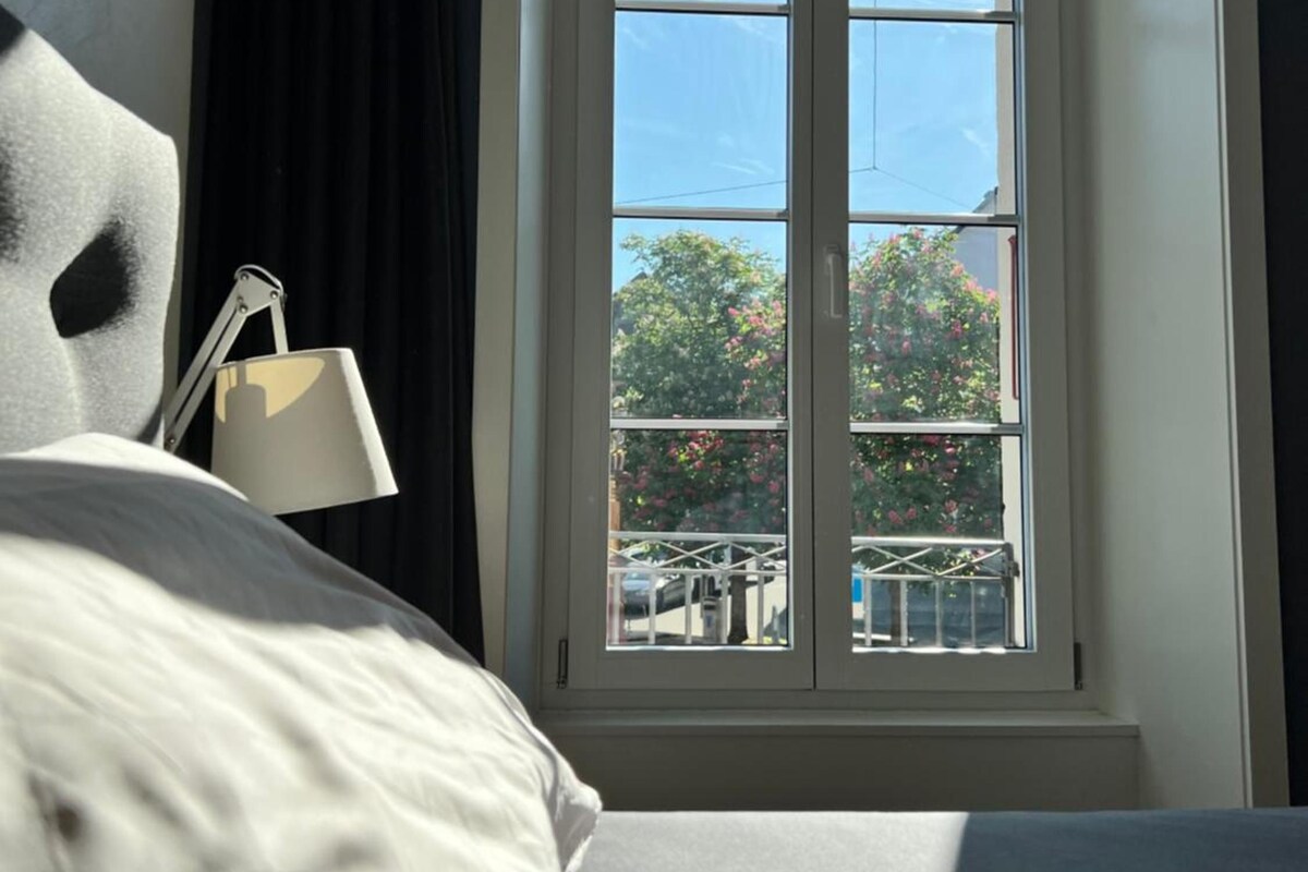 Une chambre avec une grande fenêtre, un lit, une lampe et une vue sur les arbres et un balcon.
