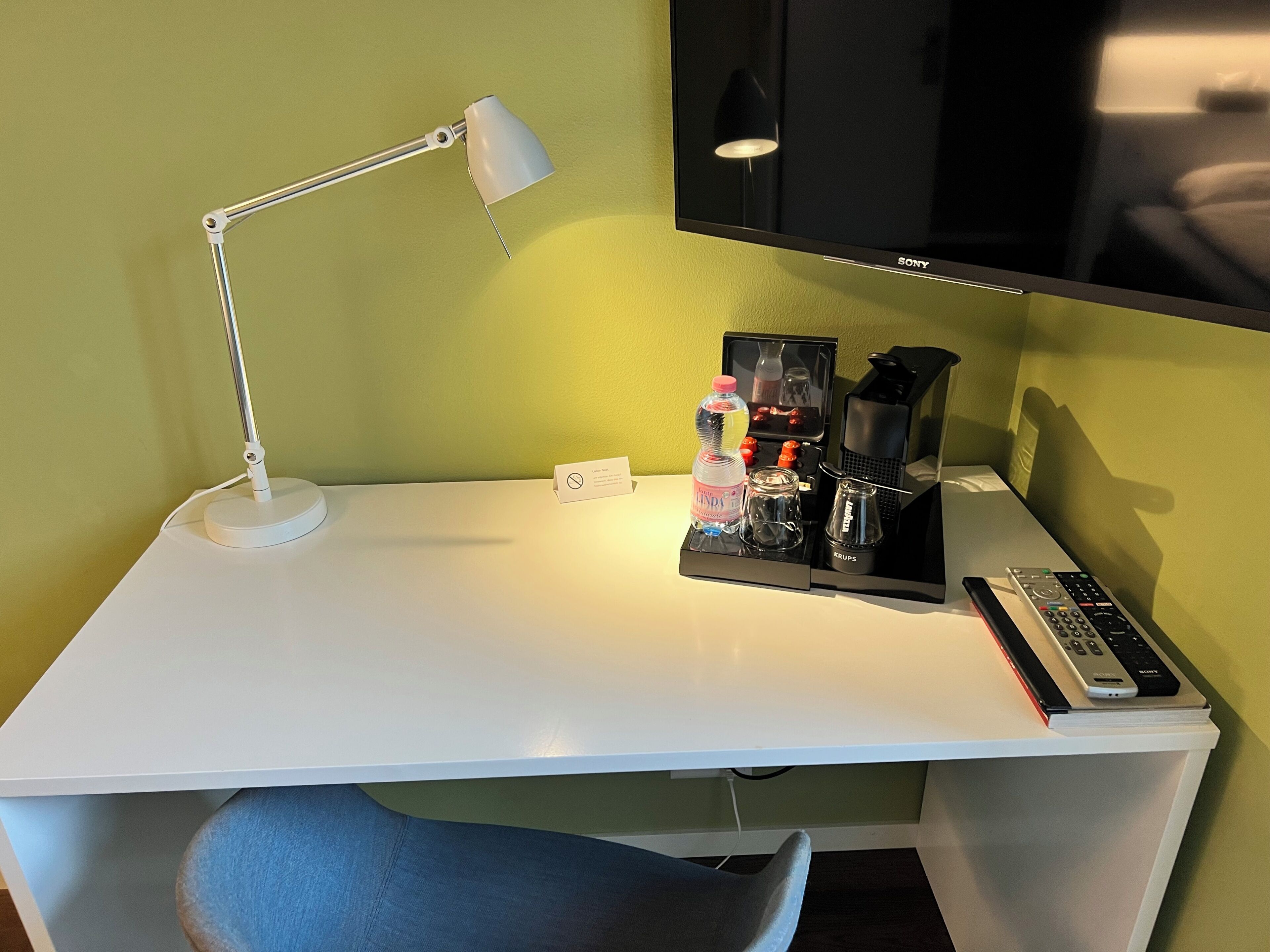 A white desk with a desk lamp, a bottle of water, a glass jar, a coffee maker, and a remote control.