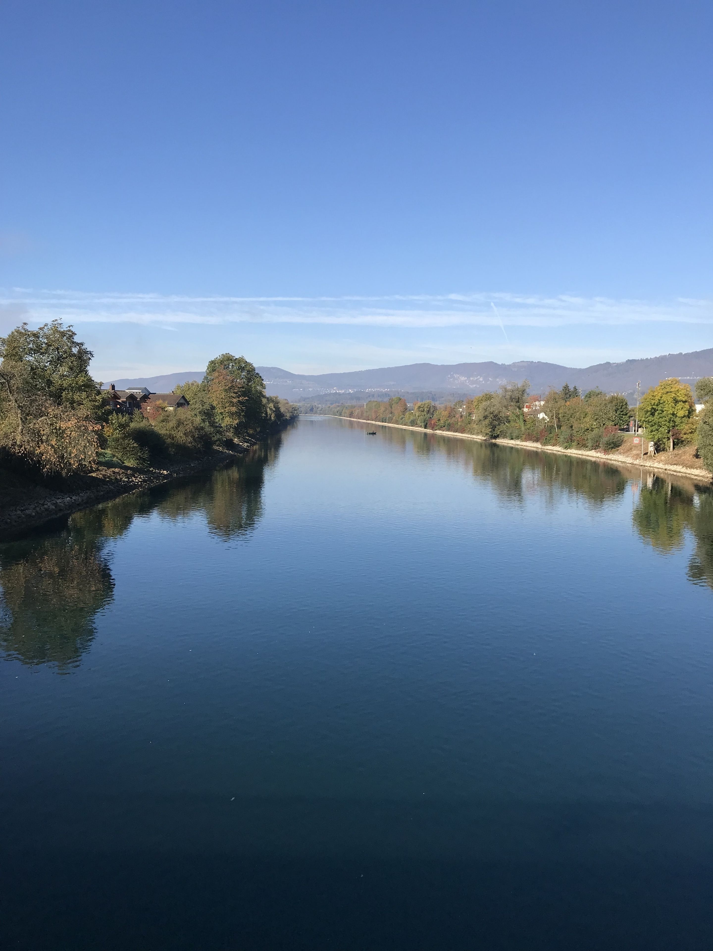 A calm river with a clear sky, surrounded by trees and distant mountains.