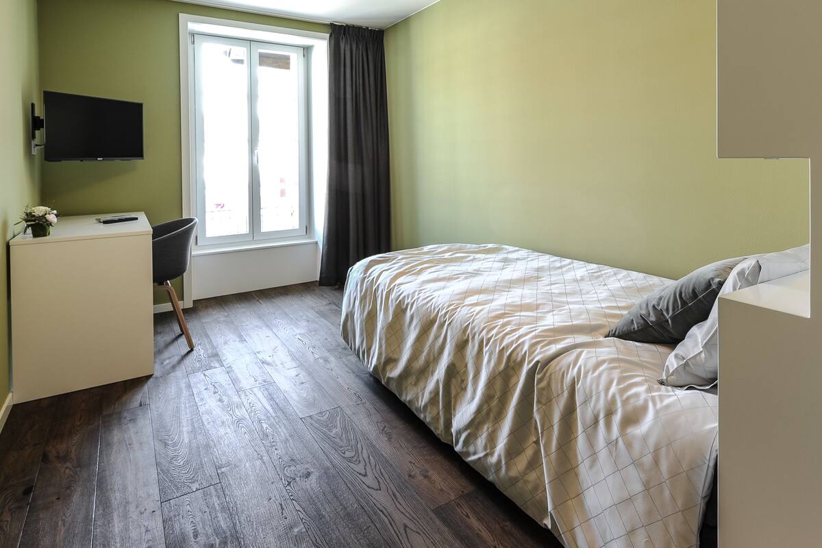 Ein Schlafzimmer mit einem Bett, einem Schreibtisch, einem Stuhl, einem Fernseher und einem Fenster mit Vorhängen.