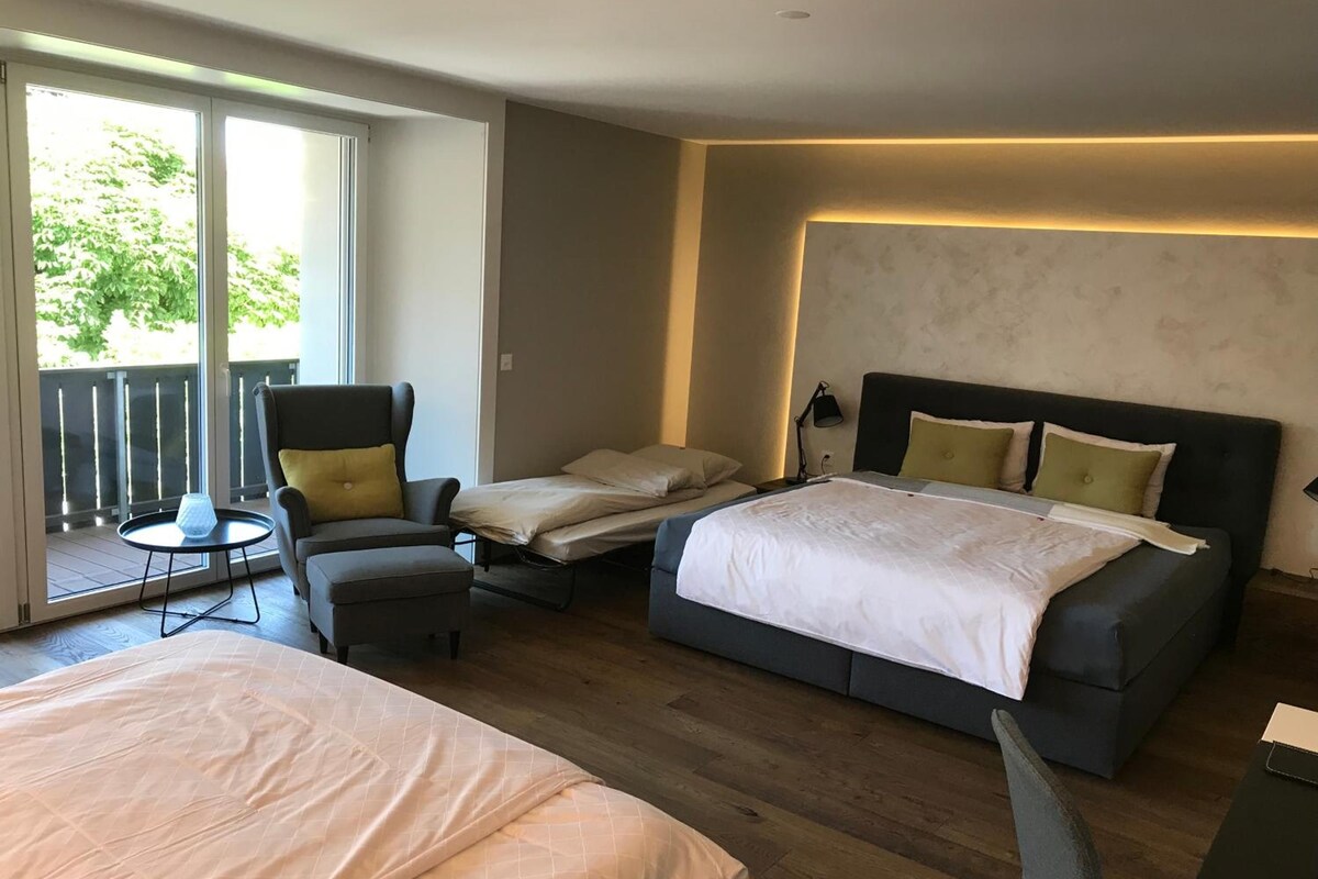 Een hotelkamer met een groot bed, een kleiner bed, een stoel, een tafel en een balkon met uitzicht op groen.