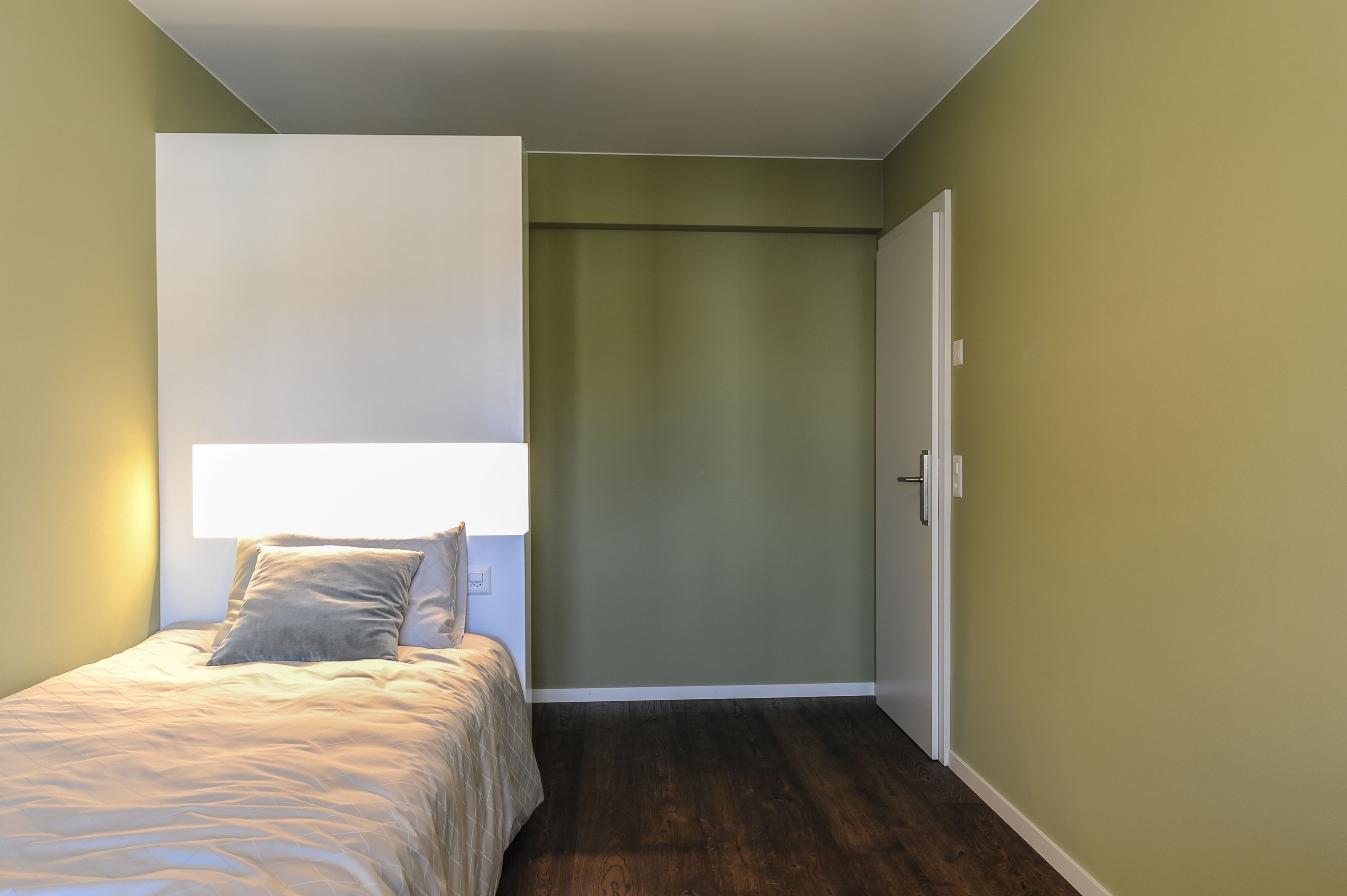 A bedroom with a bed, a nightstand, a wall-mounted light, and a door.