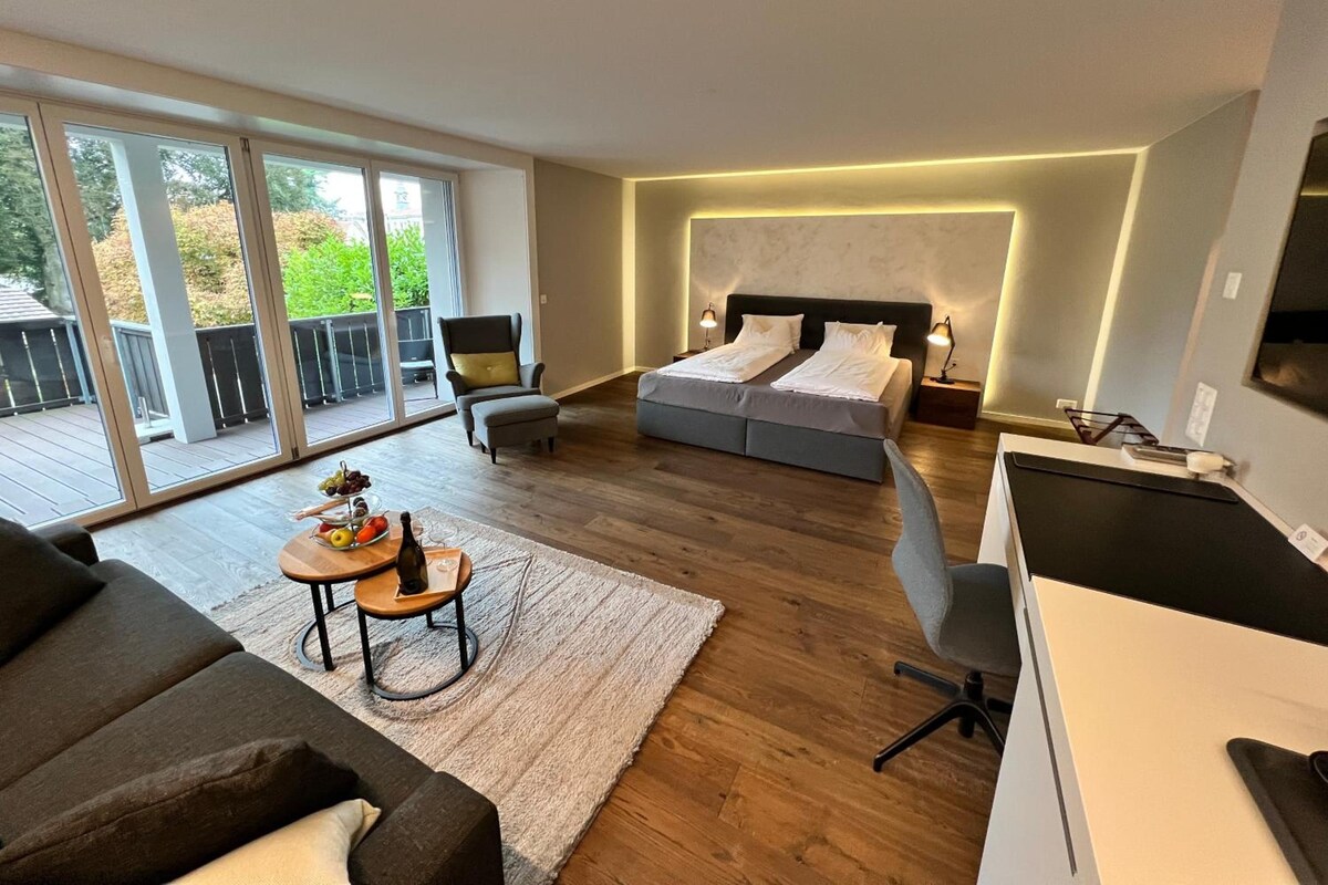 Een moderne slaapkamer met een groot bed, een bureau en een bank. Er is een balkon met uitzicht op groen.