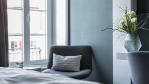 Ein Hotelzimmer mit einem Bett, einem grauen Sessel, einem kleinen Tisch, einer Vase mit Blumen und einem Fenster mit Blick auf Gebäude.