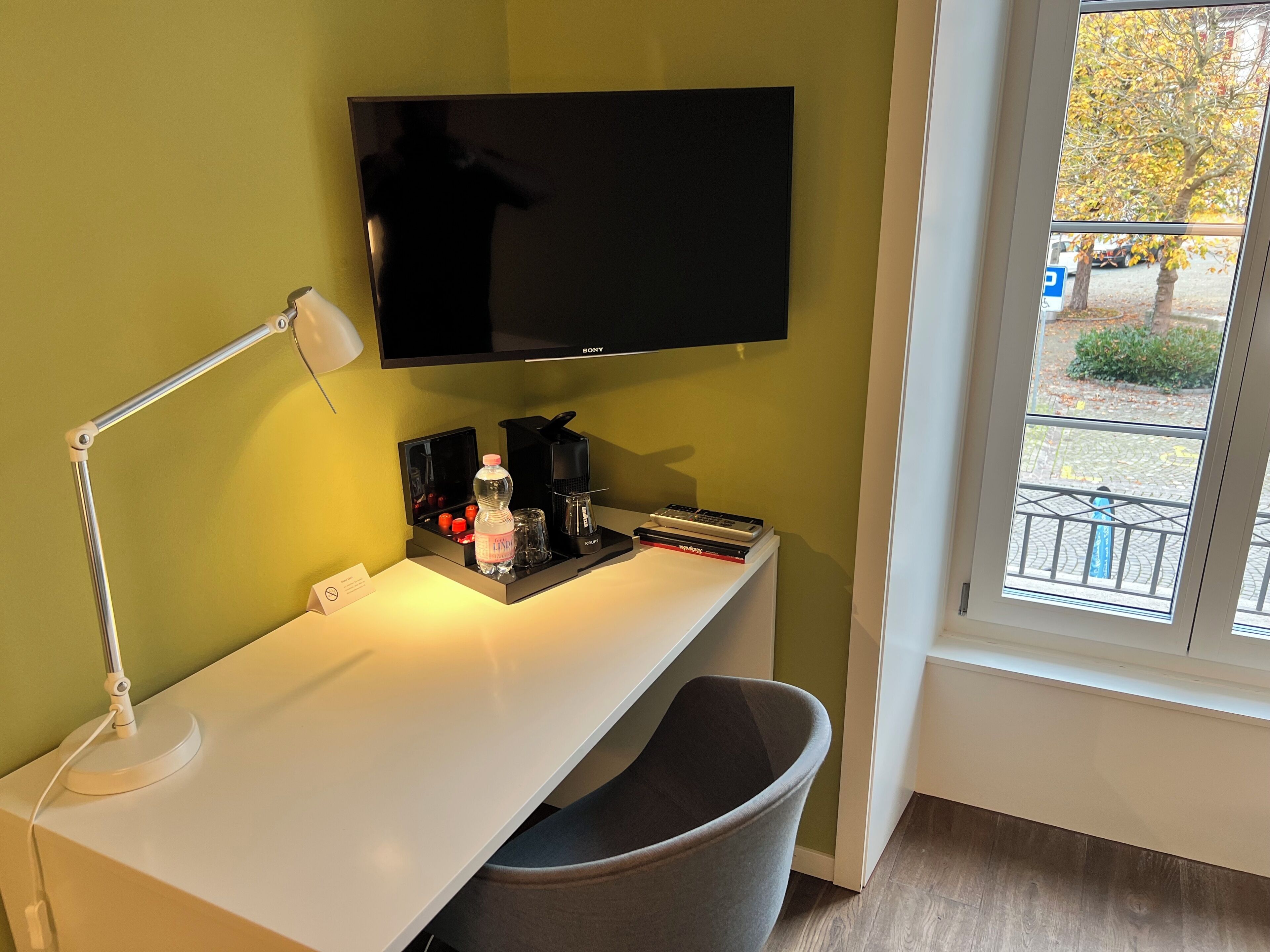 Habitación de hotel con escritorio, televisor de pantalla plana, lámpara, cafetera, cesto de basura y ventana con vistas al exterior.