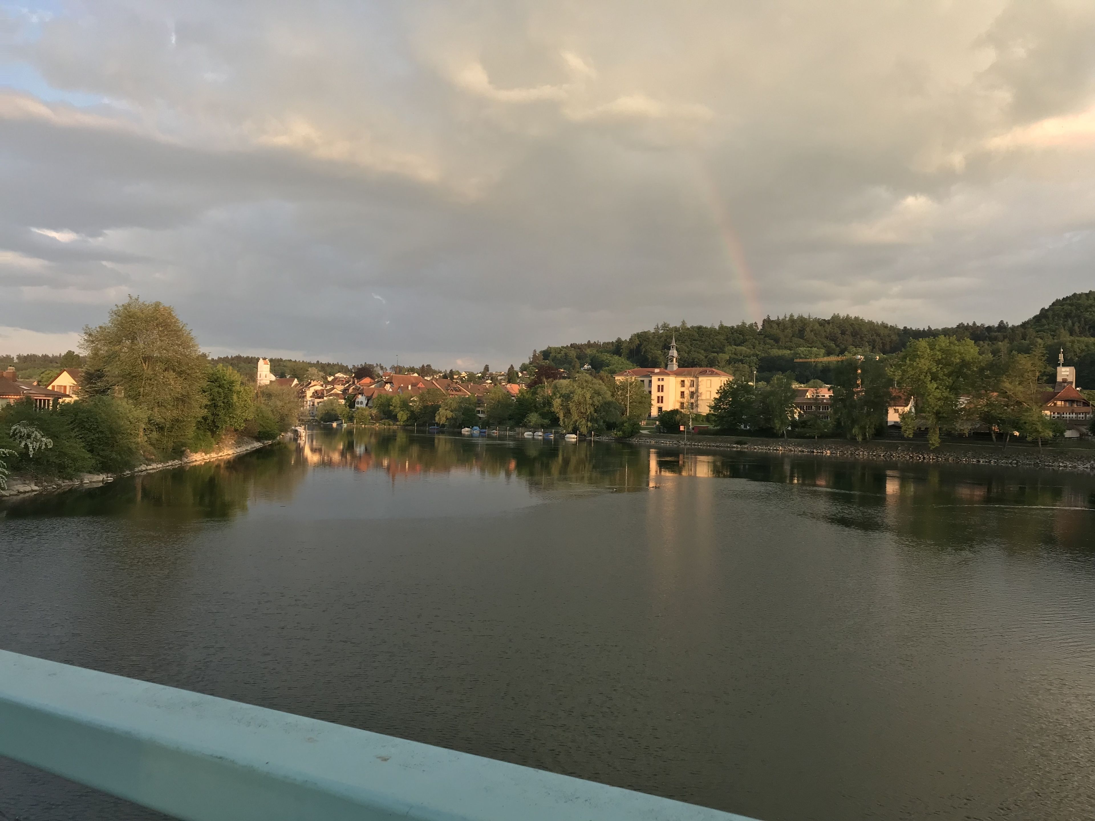 A calm river with buildings on the opposite bank, a rainbow in the sky, and a forested hill in the background.