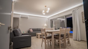 A modern living room with a grey sofa, wooden dining table, and chairs. There is a large window with curtains and a view of the outdoors. - Zante Suites (Zakynthos)