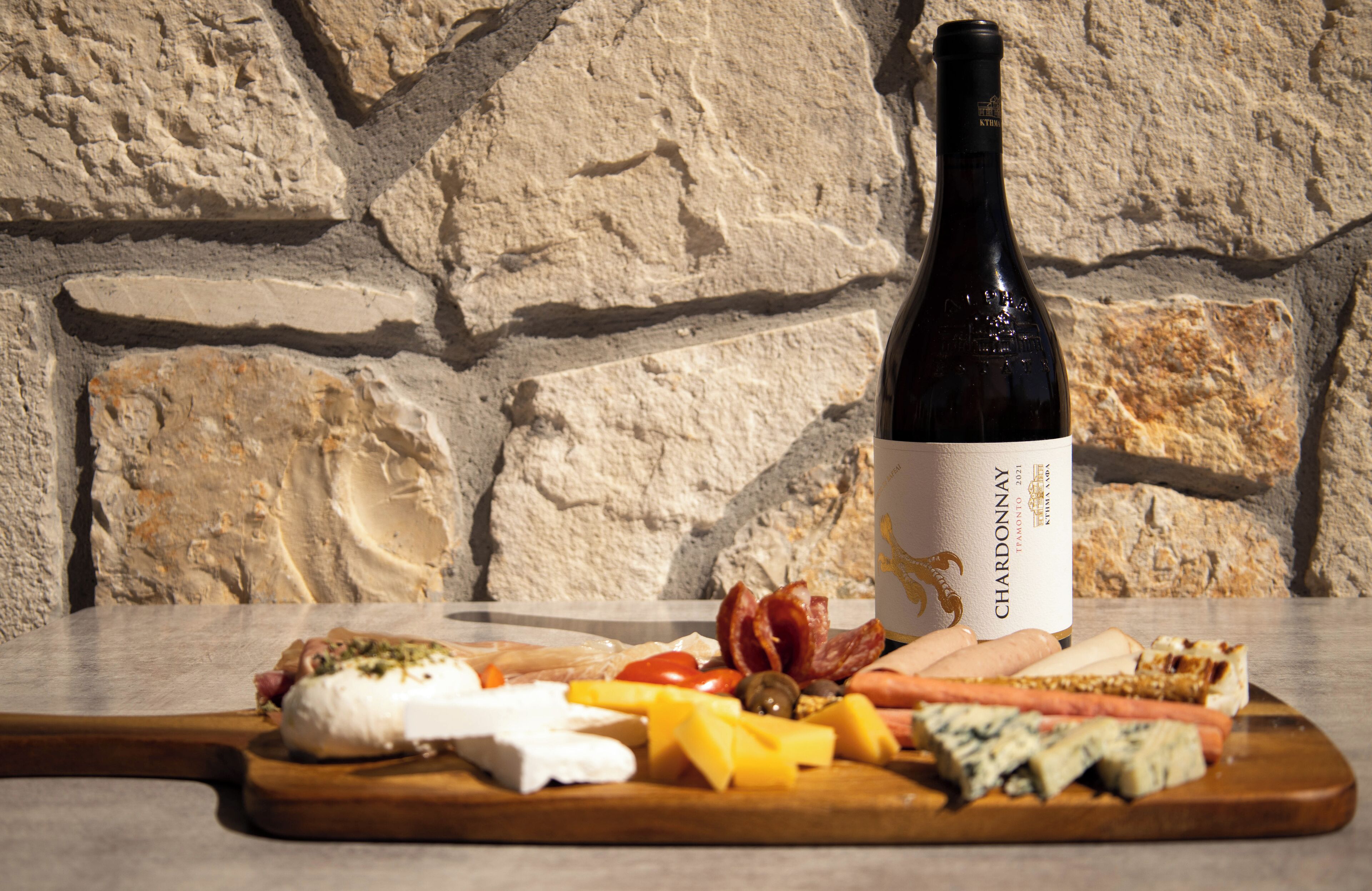 A bottle of Chardonnay wine, a wooden cutting board with various cheeses, meats, and vegetables.