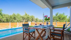 A wooden deck with a pool, outdoor table set for dining, and a view of a wooden fence and trees. - Zante Suites (Zakynthos)