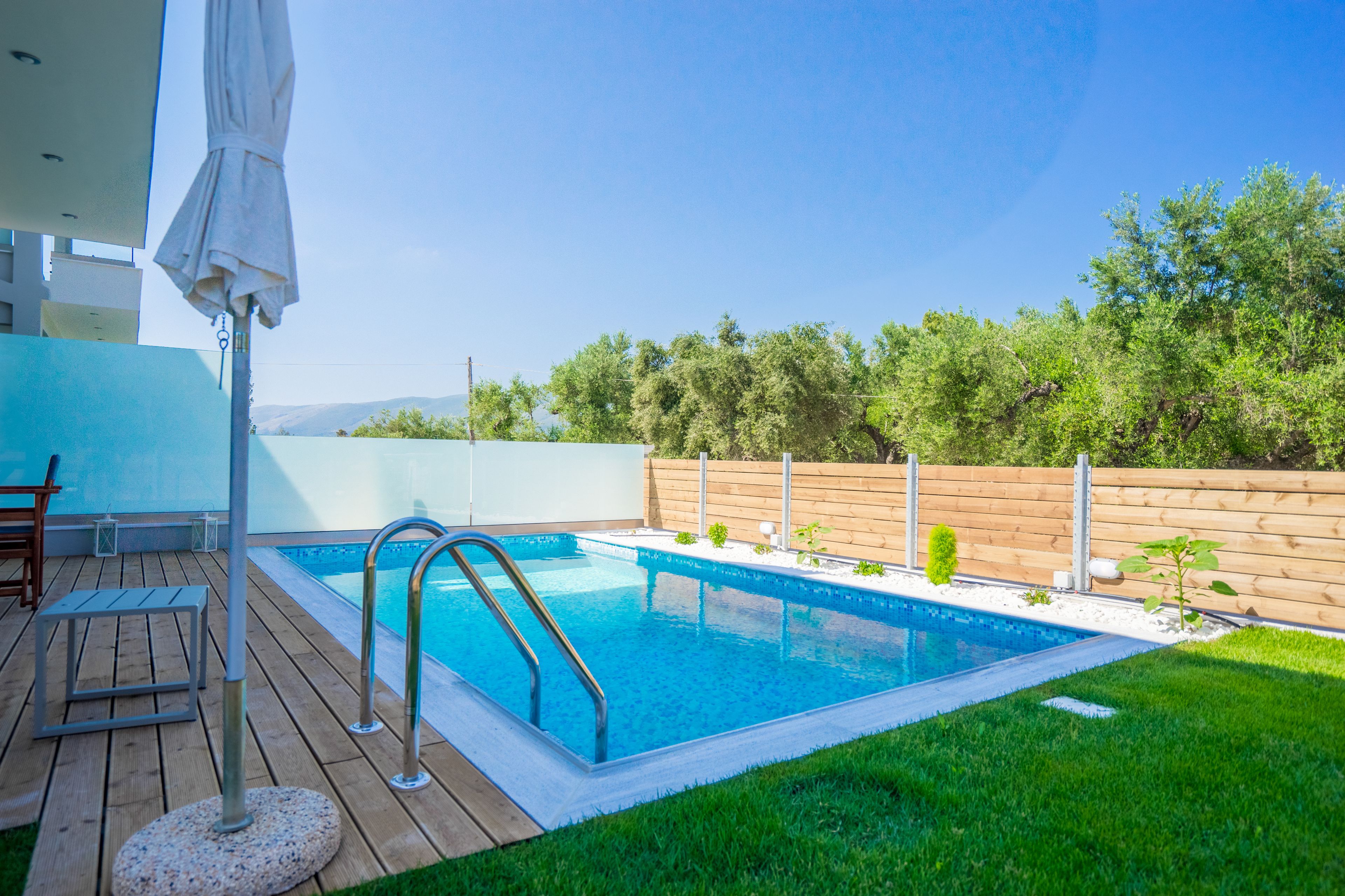 Une piscine avec une terrasse en bois, un escalier en métal et un parasol blanc.