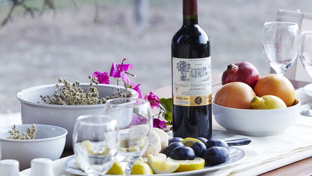 Une table dressée avec une bouteille de vin, des corbeilles de fruits et des verres, avec en arrière-plan une vue sur des arbres.
