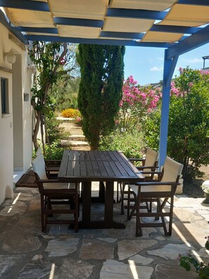 An outdoor patio with a wooden table and chairs, stone flooring, and a view of a garden with various plants and flowers.