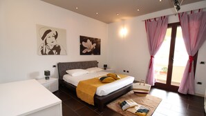 A bedroom with a bed, bedside tables, a nightstand, a bedside lamp, a window with curtains, and artwork on the wall.