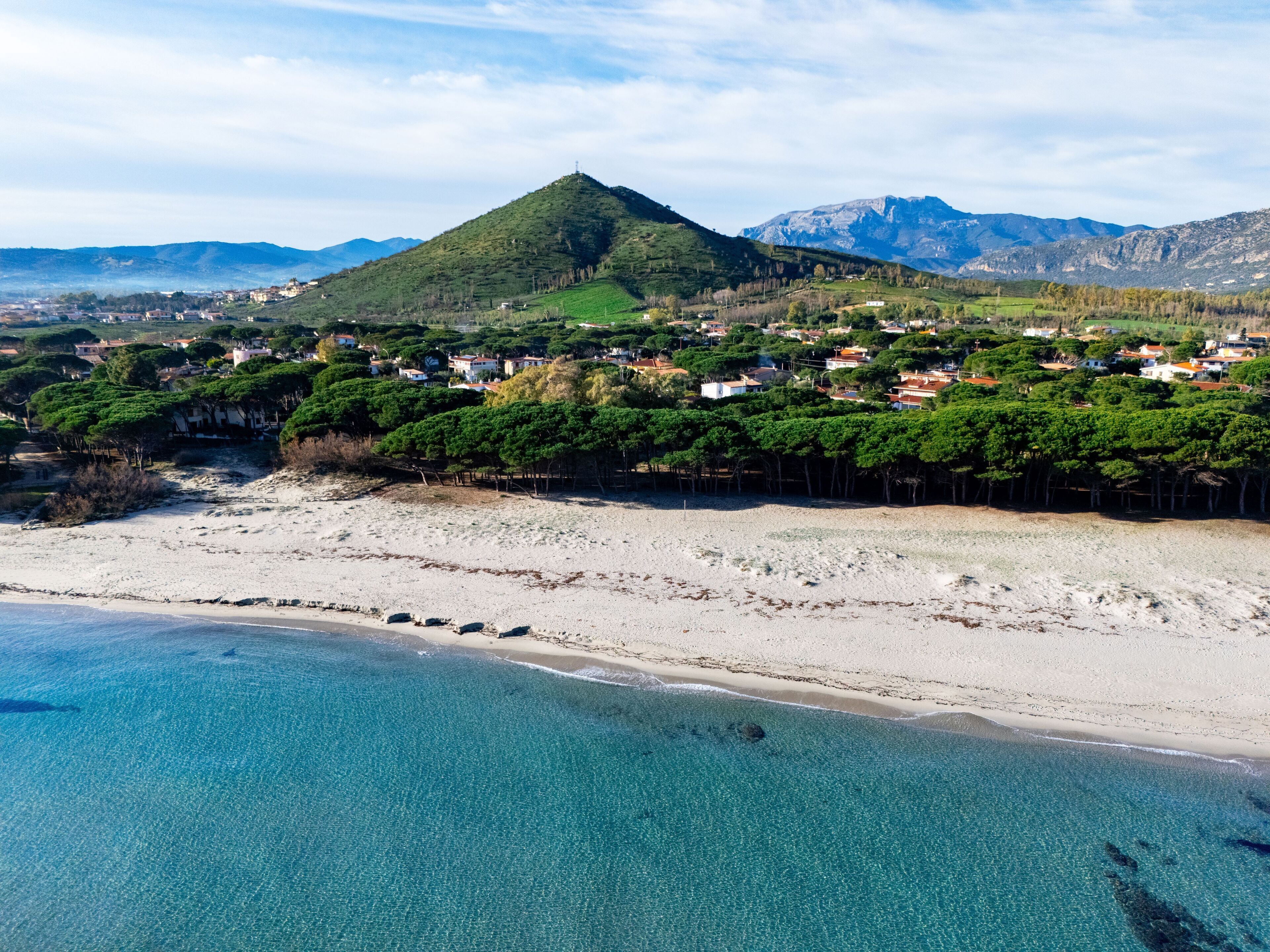 Vue aérienne d’une zone côtière avec une plage, des eaux turquoise et limpides, et une ville avec des maisons et de la végétation.
