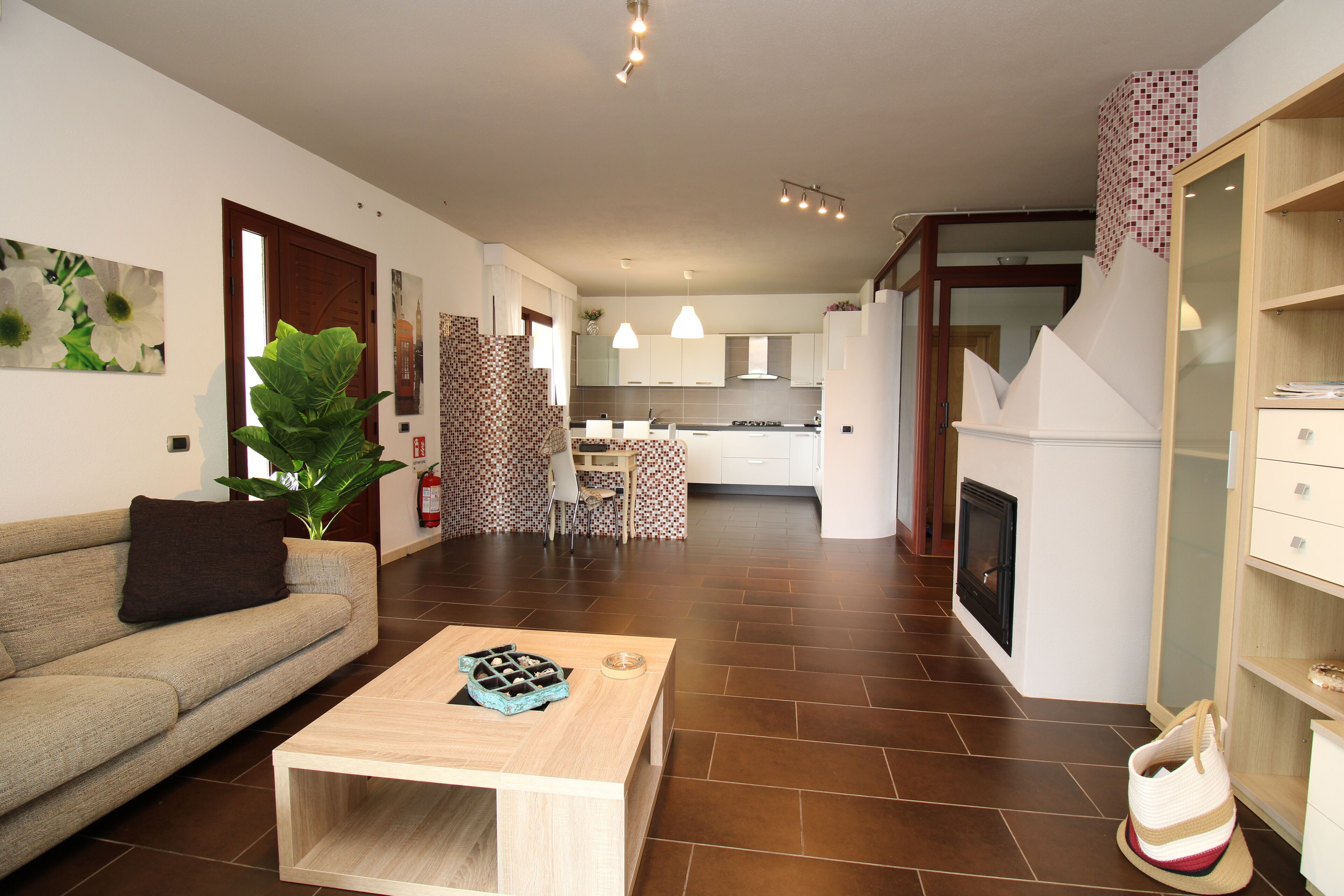 Ein modernes Wohnzimmer mit einer Couch, einem Couchtisch und einer großen Pflanze. Im Hintergrund ist eine Küche mit weißen Schränken und einem Essbereich zu sehen.