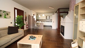 A modern living room with a sofa, coffee table, and a large plant. A kitchen with white cabinets and a dining area is visible in the background.