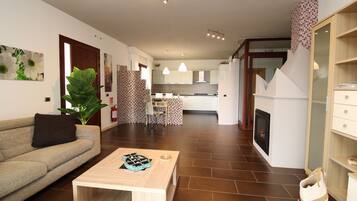 Ein modernes Wohnzimmer mit einer Couch, einem Couchtisch und einer großen Pflanze. Im Hintergrund ist eine Küche mit weißen Schränken und einem Essbereich zu sehen.