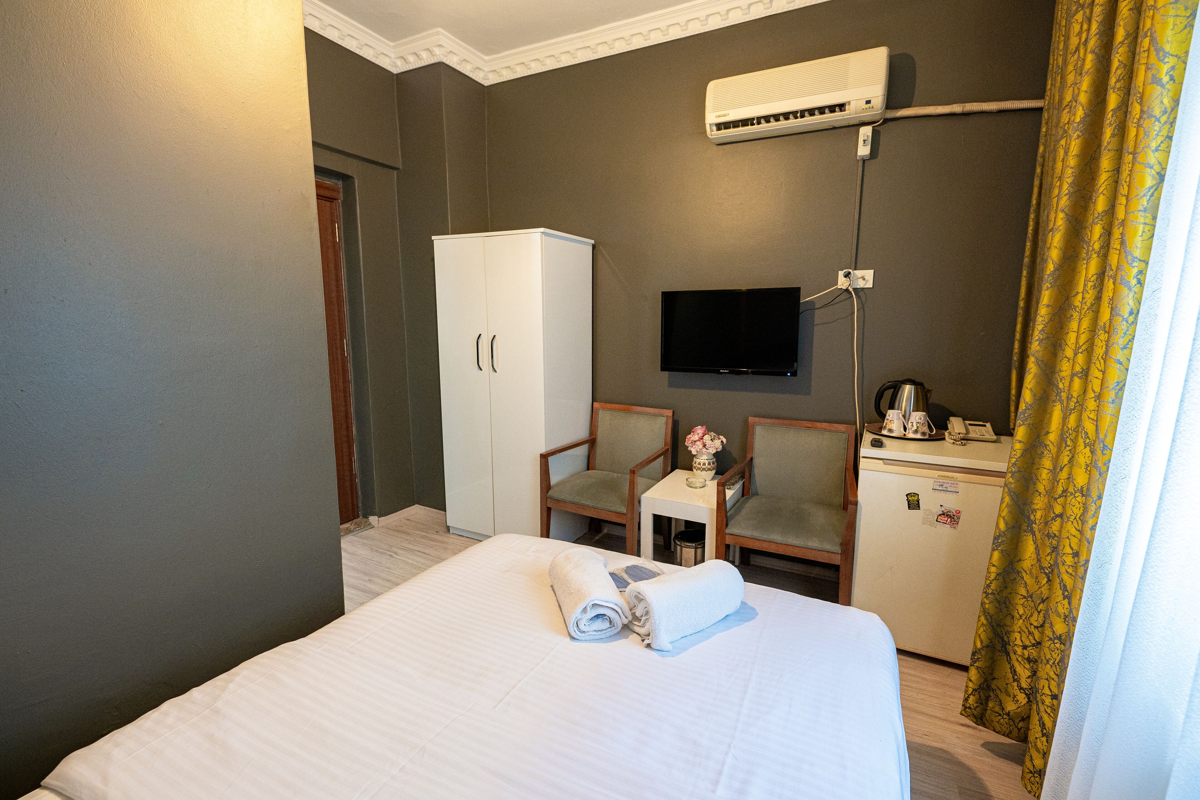 A hotel room with a bed, a desk with a chair, a television, a minibar, and an air conditioning unit.