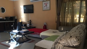 A living room with a sofa, a coffee table with a laptop, a TV mounted on the wall, and a bed with a red blanket. - Blomsbed Inn (Palapye)