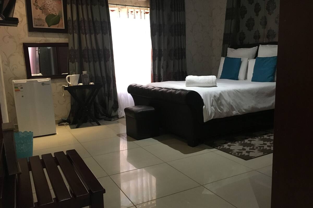 A hotel room with a bed, a small table, a bench, a refrigerator, and a window with curtains.