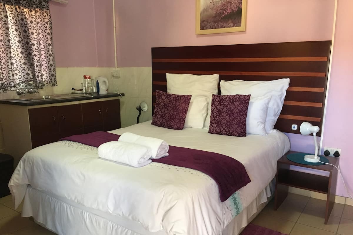 A neatly made bed with white and purple bedding, a bedside table with a lamp, and a small cabinet with toiletries.