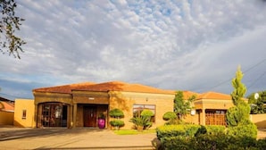 A two-story building with a tiled roof, a brick facade, and a small front garden with a purple pot and trimmed bushes. - Blomsbed Inn (Palapye)