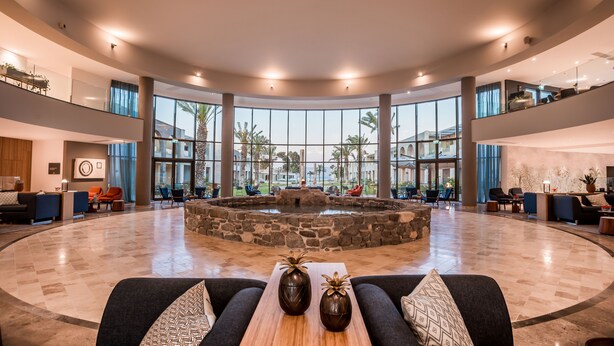 A spacious lobby with a central stone fountain, multiple seating areas, and large windows offering views of palm trees.