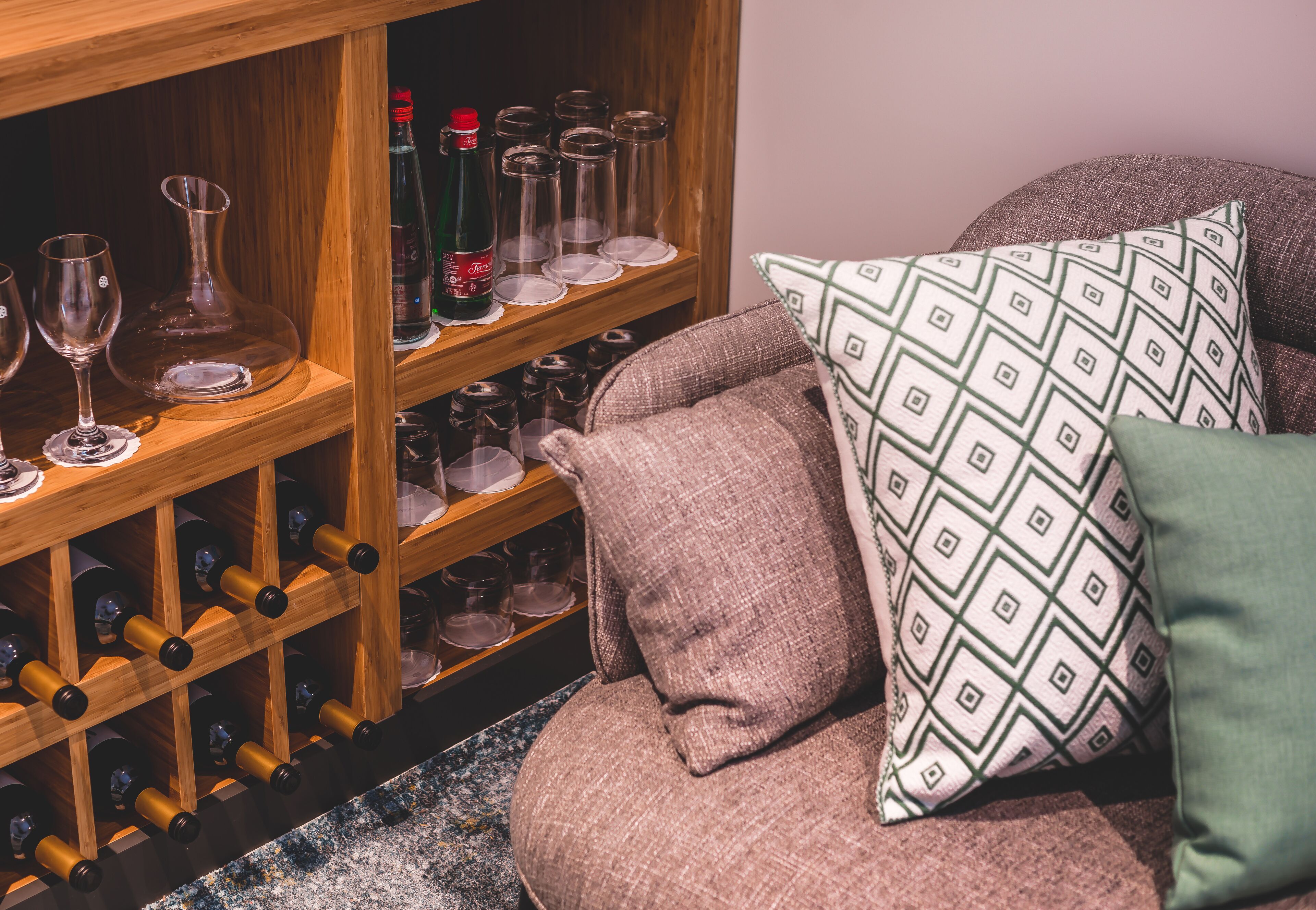 Una botellera con botellas y copas, un sofá acolchado con cojines estampados y un estante de madera.