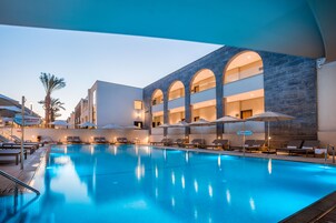 Una piscina con sillas de descanso y sombrillas, un edificio con ventanales arqueados y una palmera.