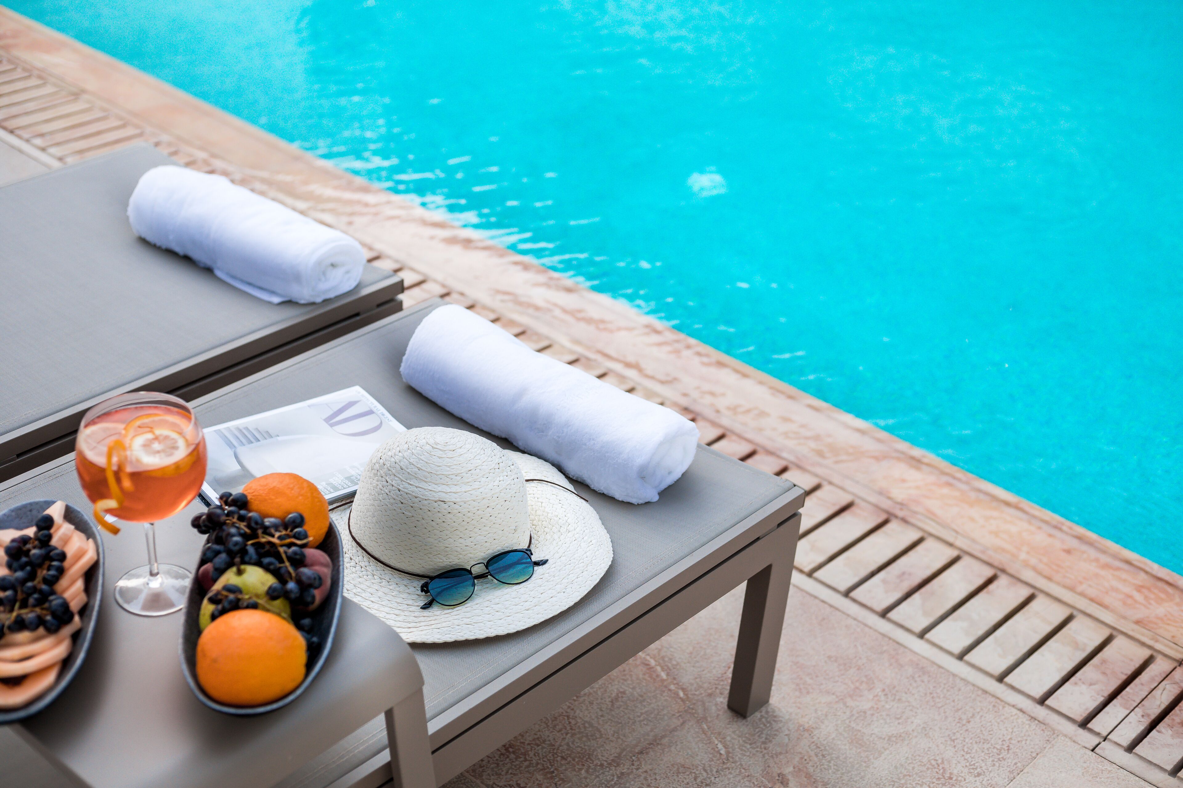 Mesa junto a la piscina con una bebida, frutas, un sombrero y toallas.