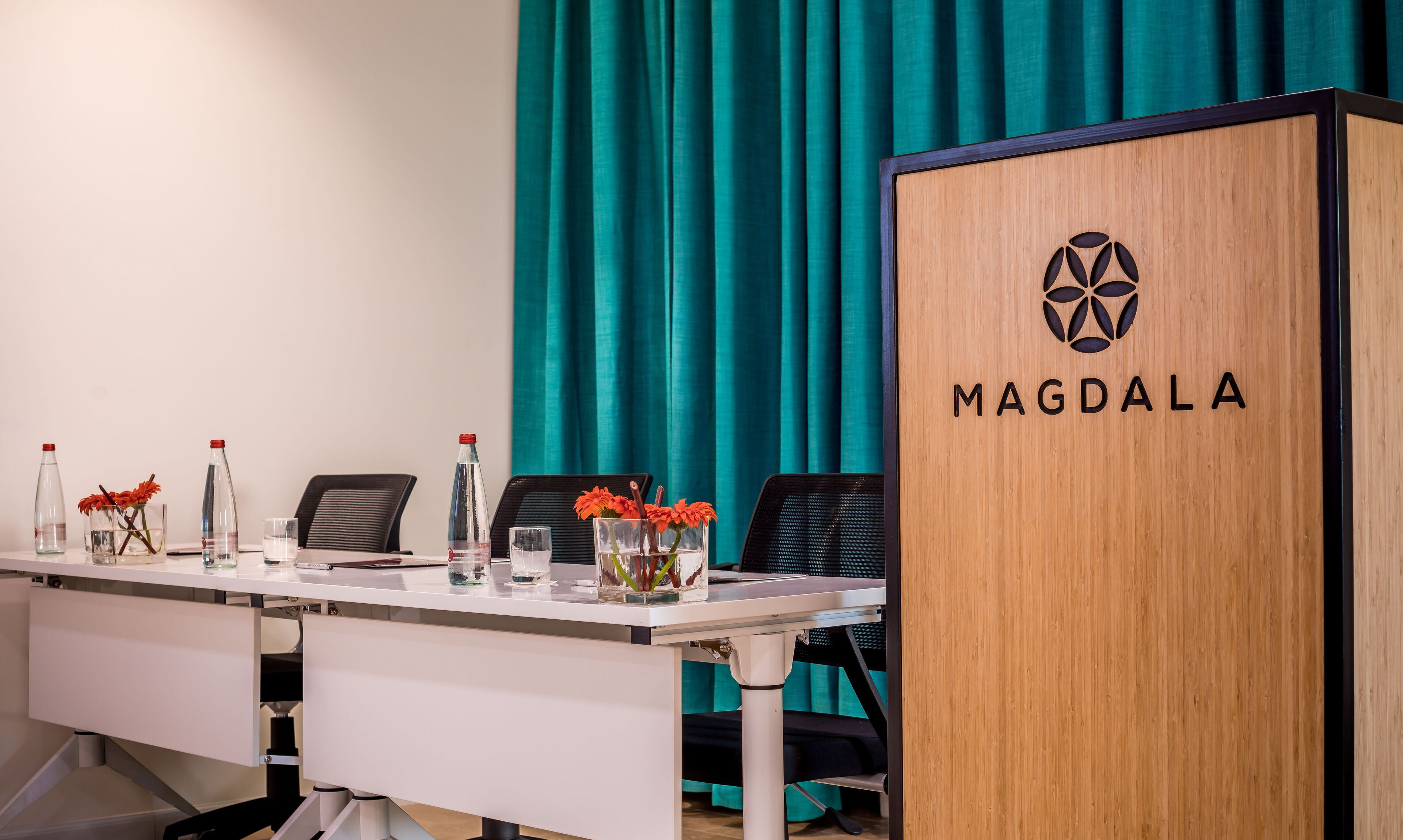 Un salón de conferencias con un atril de madera con la etiqueta "MAGDALA", una mesa blanca con sillas y cortinas color turquesa al fondo.