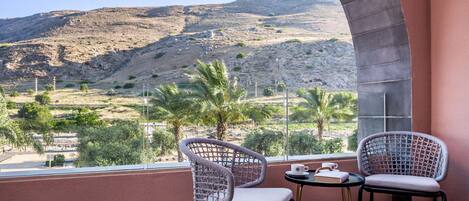A balcony with two wicker chairs, a small table, and a view of a hilly landscape with sparse vegetation.