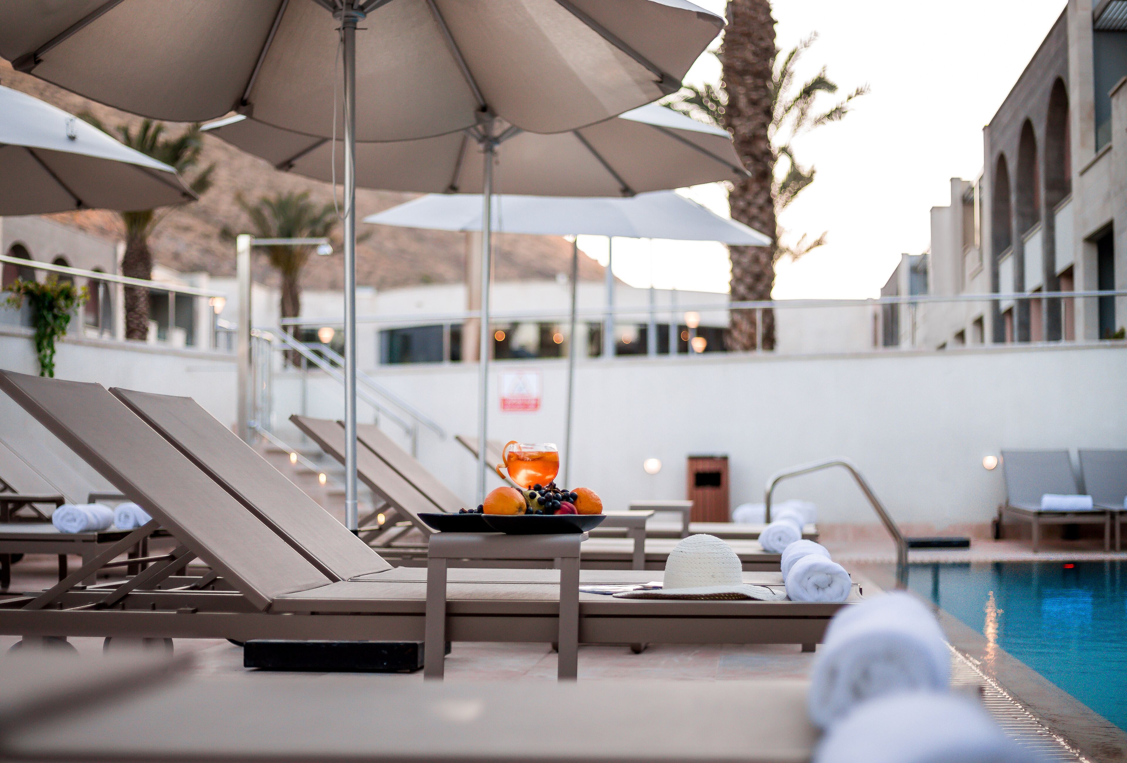 A poolside area with lounge chairs, umbrellas, and a table with fruit and a hat.