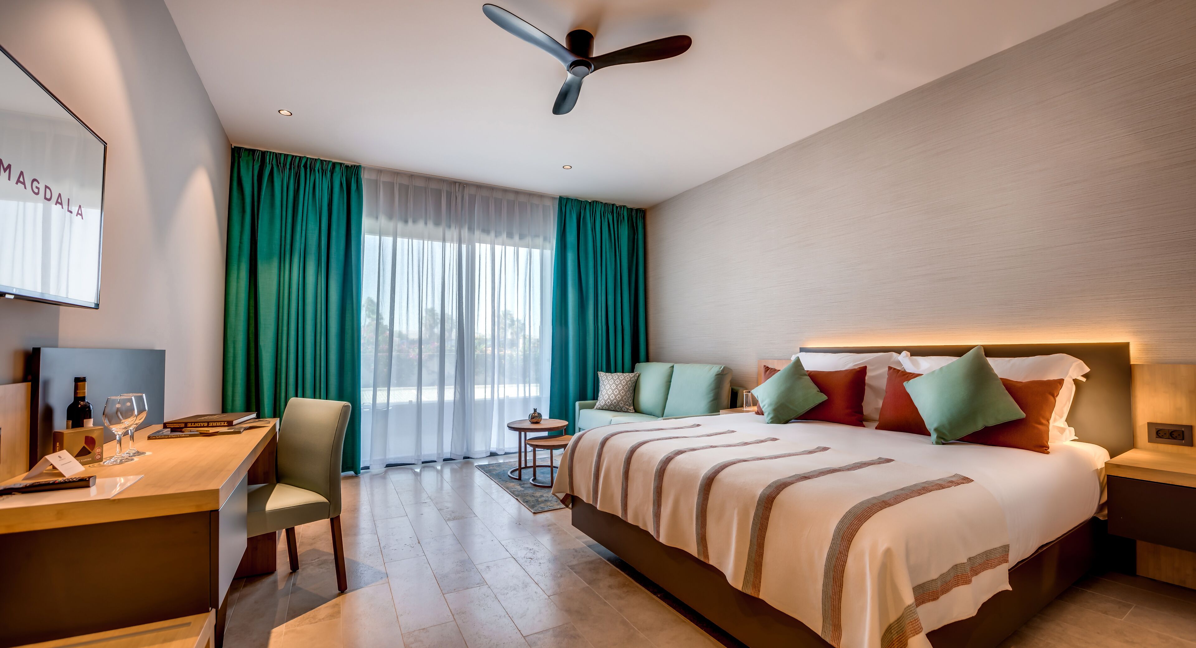 A hotel room with a bed, a desk with a chair, a small table, and a ceiling fan.