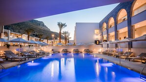 A swimming pool with lounge chairs and umbrellas, surrounded by buildings with arched windows and a mountainous backdrop.