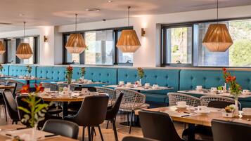 A modern dining area with blue upholstered booths, wicker chairs, and hanging pendant lights. Tables are set with white tableware and vases with flowers.