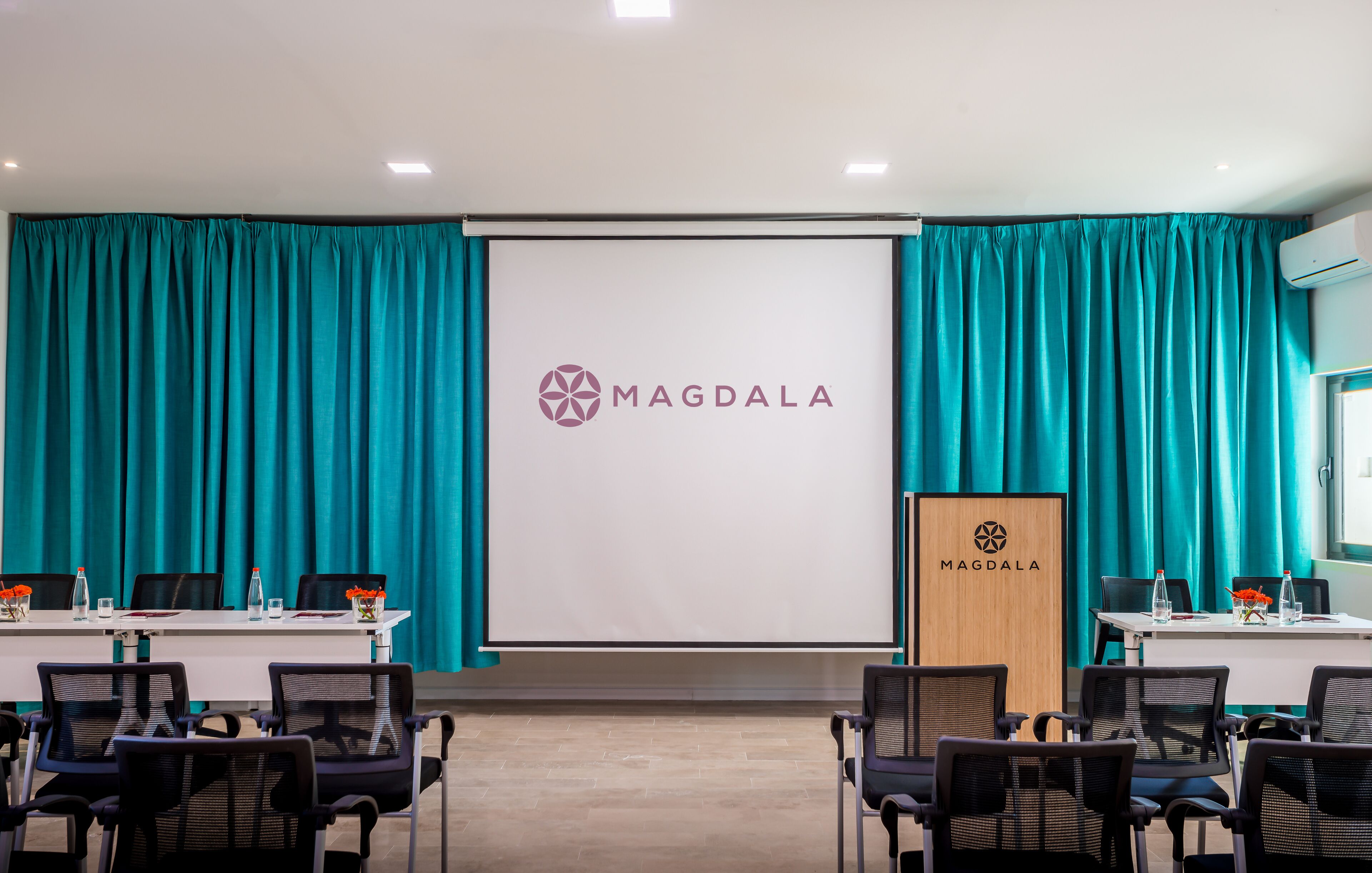 Un salón de conferencias con cortinas azul verdoso, una pantalla blanca que muestra el logo de MAGDALA y un atril de madera.