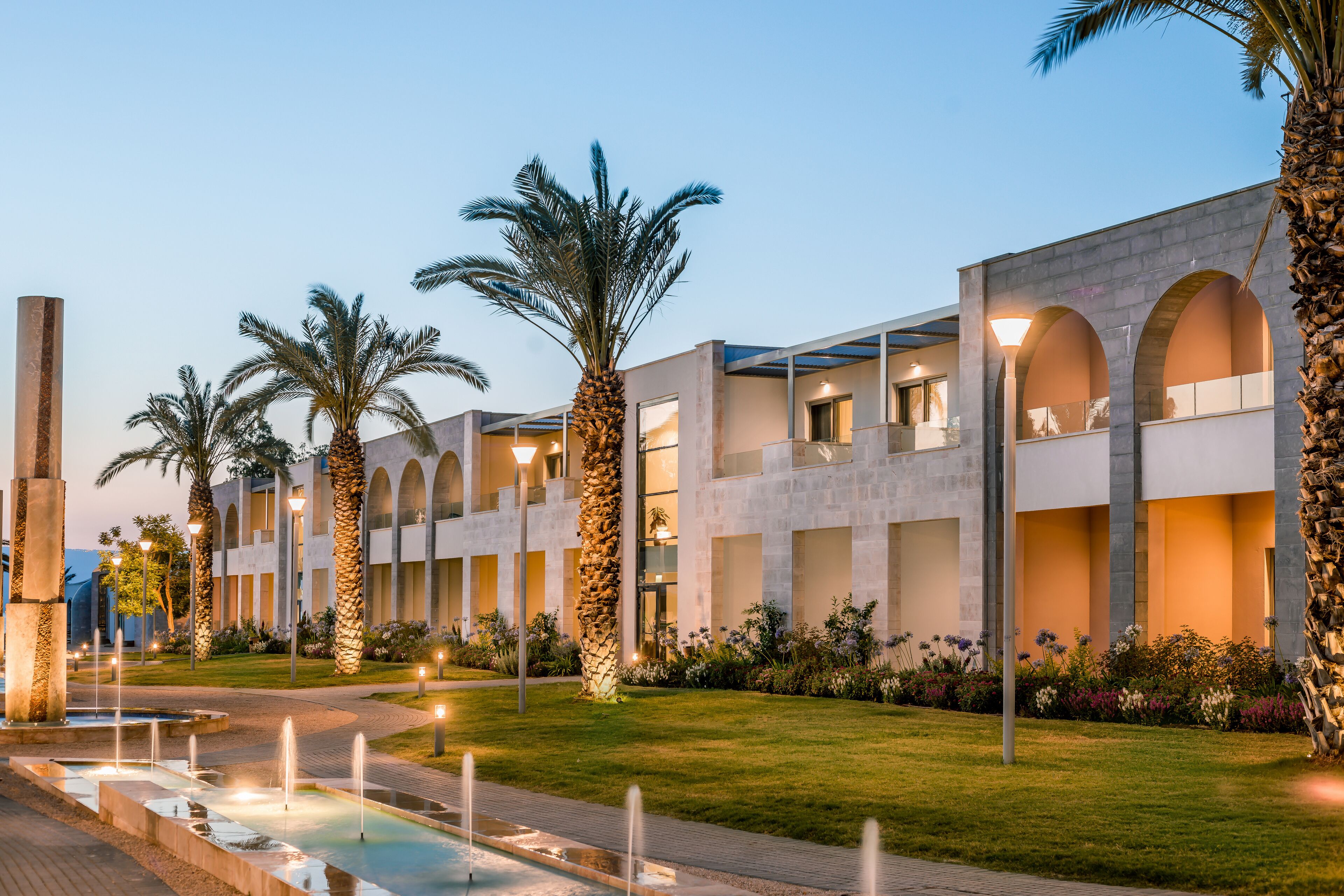 A modern building with arched windows and a well-maintained garden with palm trees and a fountain.
