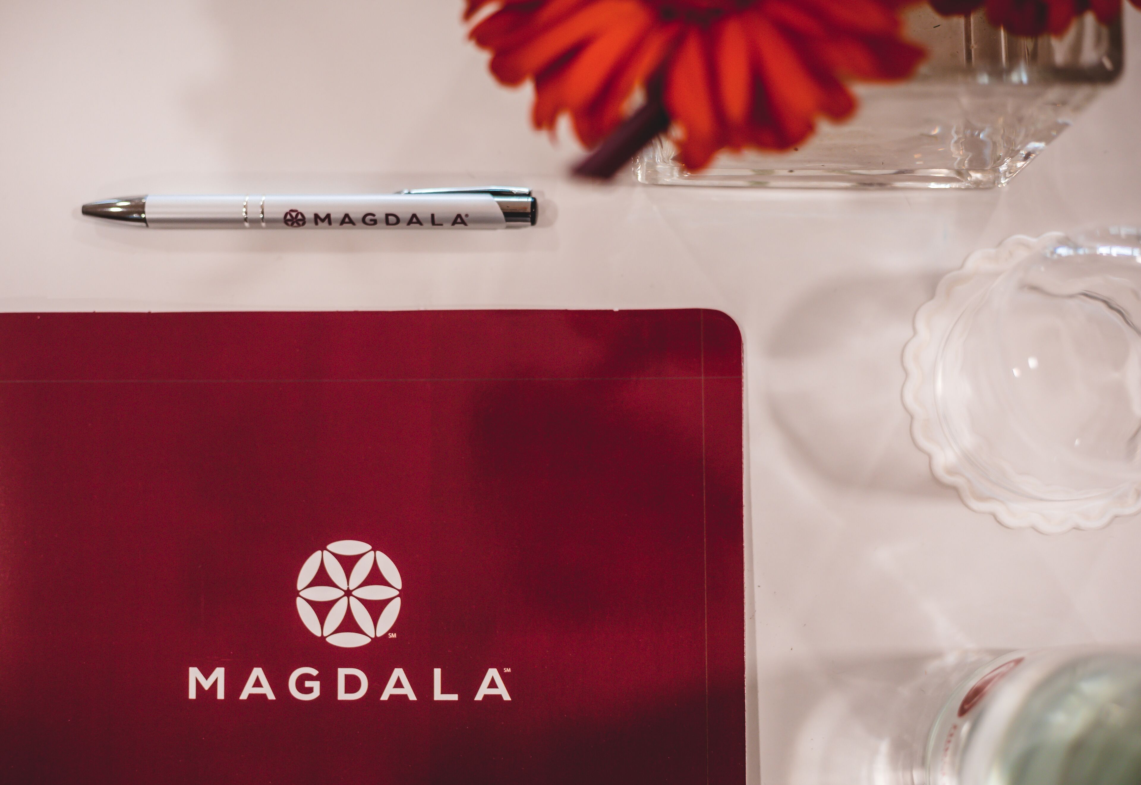 Una libreta de la marca MAGDALA con un logotipo, un bolígrafo y un florero con flores sobre una mesa.