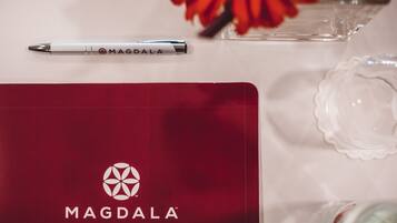 Una libreta de la marca MAGDALA con un logotipo, un bolígrafo y un florero con flores sobre una mesa.