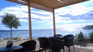 Un área de estar al aire libre techada con sillas de mimbre y una mesa, con vista a un cuerpo de agua y una costa lejana.