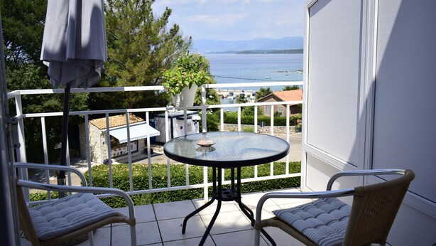 Een balkon met een ronde tafel en twee stoelen, met uitzicht op een wateroppervlak en een verre kustlijn.