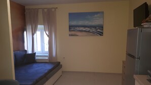 A compact living space with a bed, a small window with curtains, a painting of a beach scene, and a refrigerator. - Apartment Sabina & Grapes (Malinska-Dubasnica)