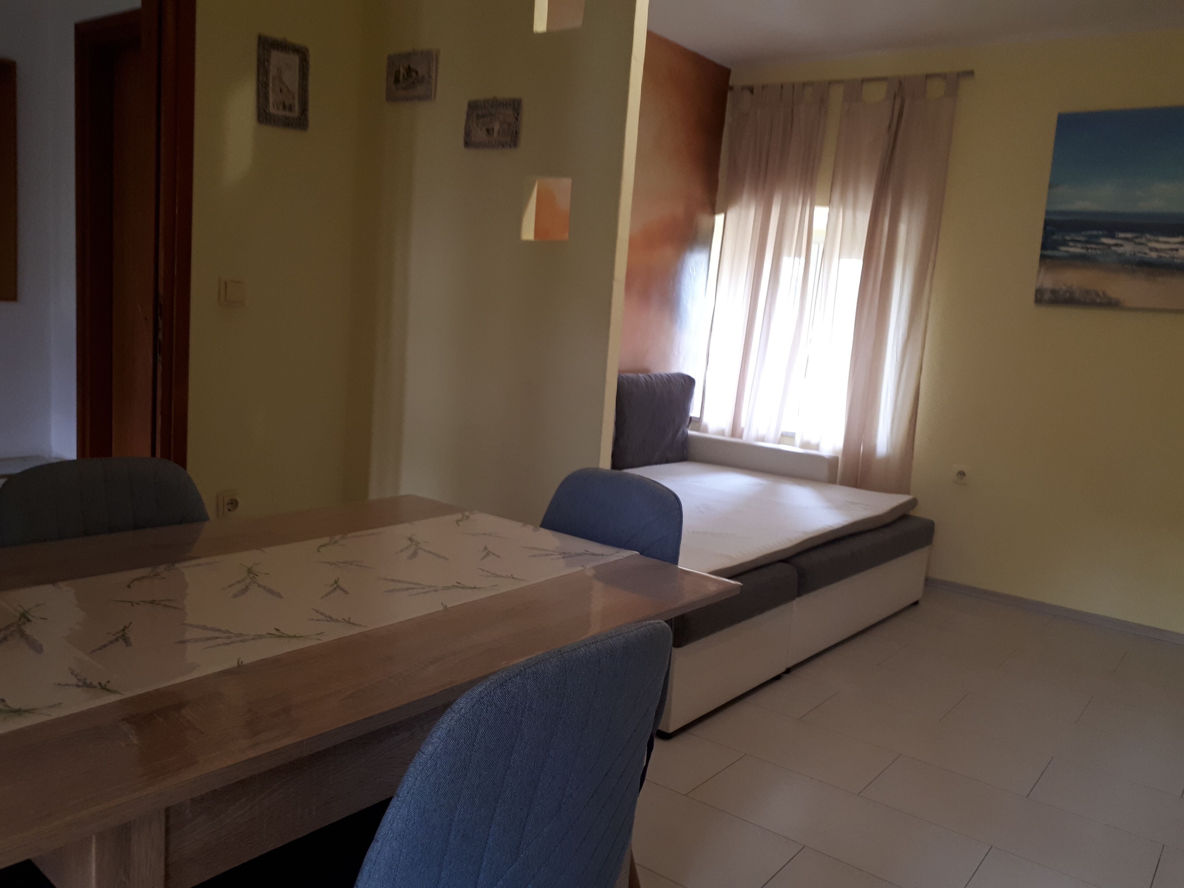 A room with a long wooden table, a sofa, a window with curtains, and a painting on the wall.