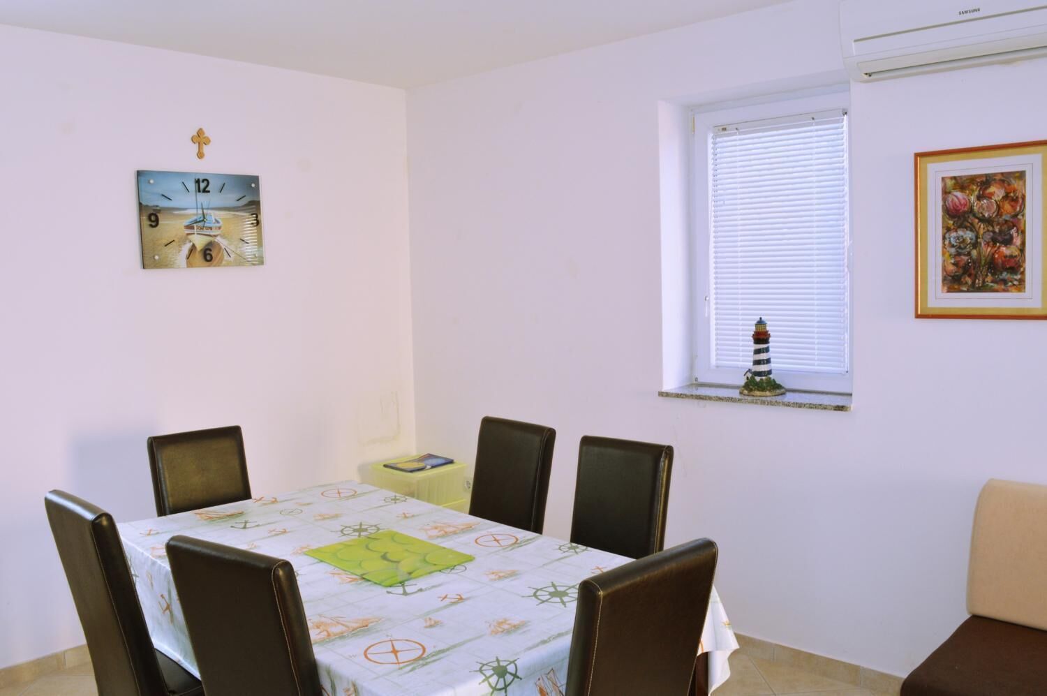 Een eetkamer met een tafel gedekt voor zes personen, een klok aan de muur, een raam met luiken en een klein beeldje op de vensterbank.