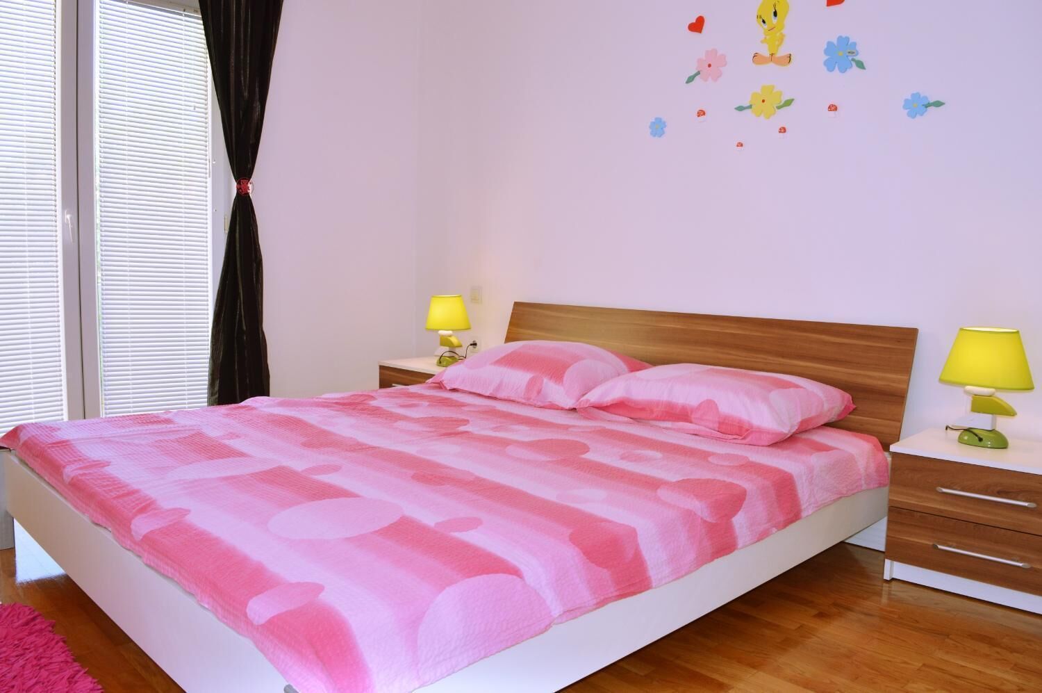 A bedroom with a bed, two bedside tables with lamps, a window with blinds, and a wall with a flower decal.