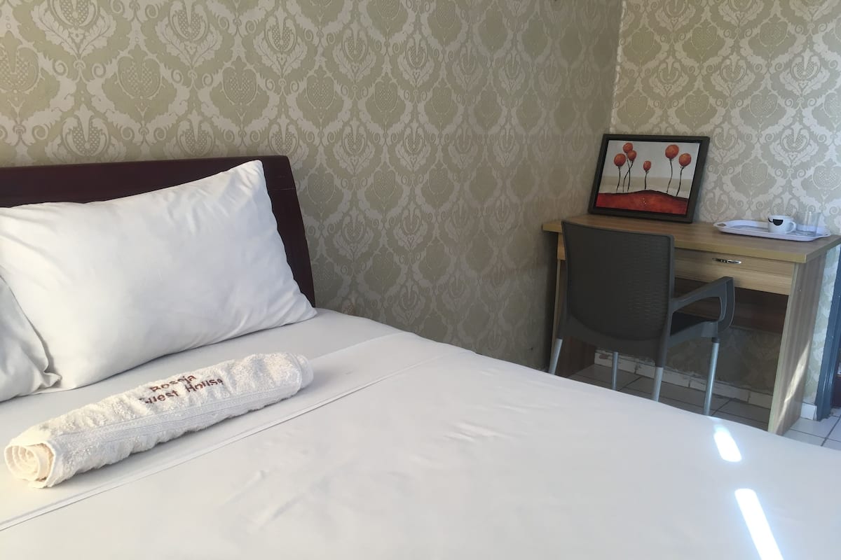 Ein ordentlich bezogenes Bett mit einem weißen Kopfkissen und einem zusammengerollten Handtuch, ein Holztisch mit einem Stuhl und ein gerahmtes Bild an der Wand.