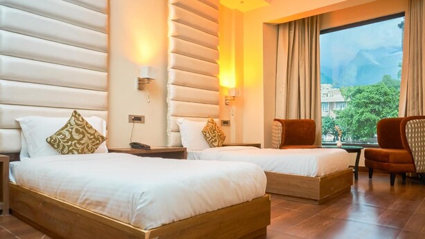 A hotel room with two beds, a view of greenery outside, and a large window with curtains. - Ganpati Hotel (Reasi)